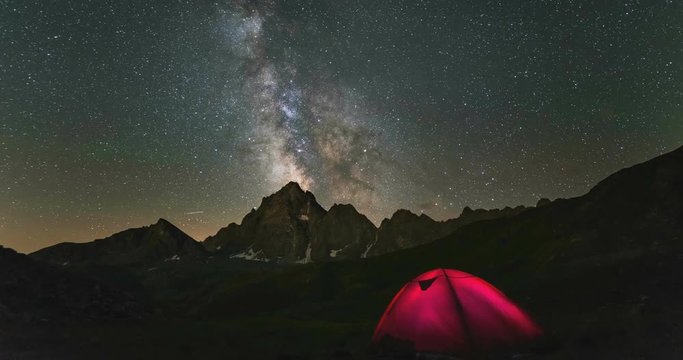 Time lapse Milky way galaxy stars rotating over the Alps, camping illuminated tent, snowcapped mountain peak, astro night sky stargazing