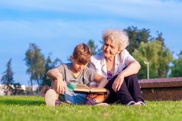 Attractive grandmother with her teenage grandson are reading a book outdoors together. Family relationships. Self-education and development