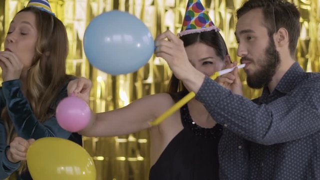 4 happy cheerful people at a party in paper caps fighting balloons. The guys are blowing in the pipes and dancing. A group of laughing people of mixed race on a gold background. HD