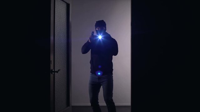 Masked man with gun and flashlight opening crime scene door, police service