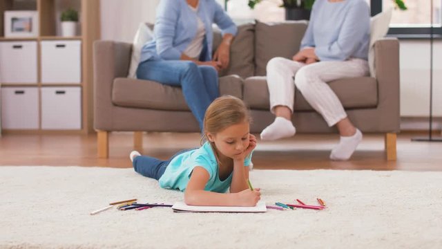 family, childhood and leisure concept - girl drawing in sketchbook by crayons lying on floor and adults sitting on sofa at home