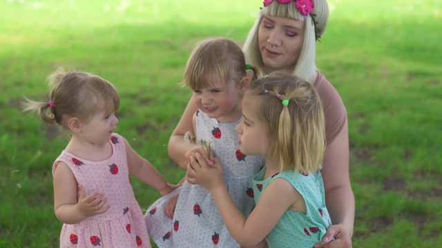 Young blonde hippie mother having quality time with her baby girls at a park blowing dandelion - Daughters wear similar dresses with strawberry print - Family values