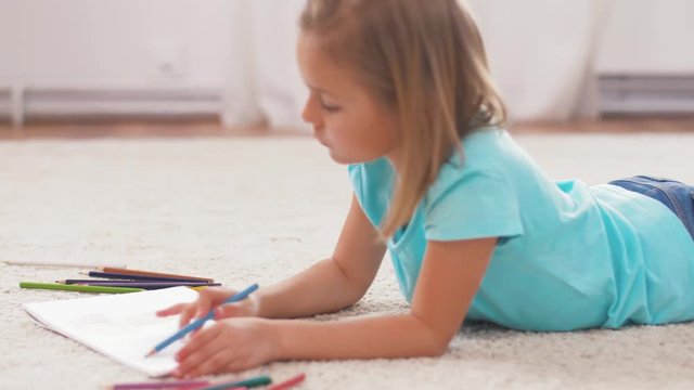 childhood, art and creativity concept - girl drawing in sketchbook by crayons lying on floor at home