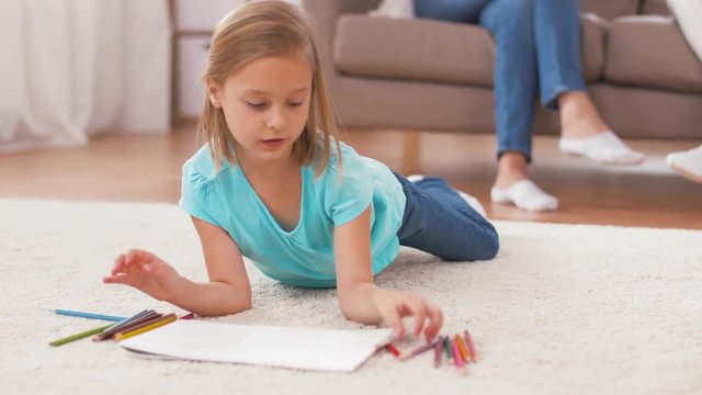 family, leisure and three generations concept - daughter drawing in sketchbook by crayons lying on floor and mother talking to grandmother on sofa at home