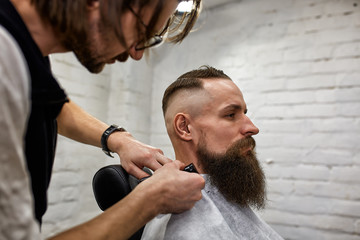 Obraz premium Master cuts hair and beard of men in the barbershop, hairdresser makes hairstyle for a young man
