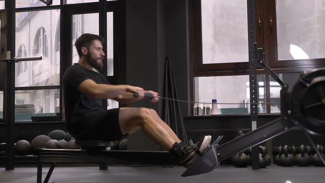 Closeup side view shoot of adult muscular athletic man using rowing machine indoors in the gym