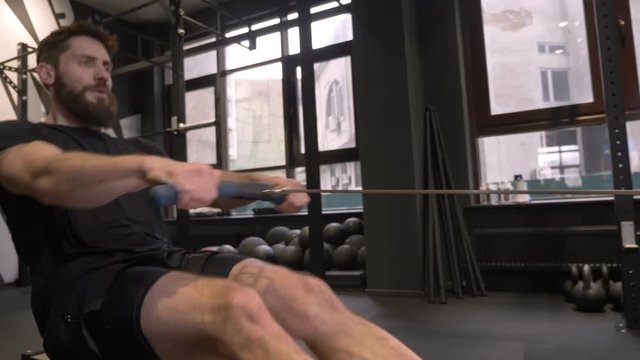 Closeup side view shoot of adult muscular athletic man using rowing machine to train indoors in the gym
