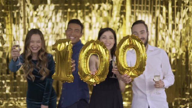 The number 100 of foil. 4 happy cheerful people at a party with balloons in their hands. A group of laughing people of mixed race on a gold background. 4K