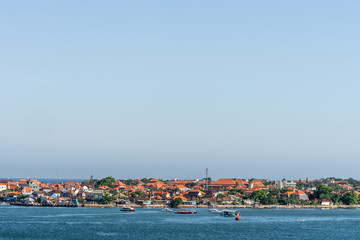 Fototapeta na wymiar Benoa harbour, Bali, Indonesia - February 26, 2019: The red roofs of the buildings on Tanyung Benoa Peninsula under light blue sky, above dark blue water, seen from harbour. Green foliage, boats.