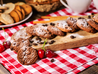 Fototapeta na wymiar Oatmeal chocolate cookies with coffee grains and cherry straws pastry with powdered sugar on kitchen cutting board and cup of tea or coffee gingham checkered fabric. Traditional dish.