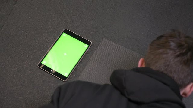 Closeup shoot of adult muscular athletic man pushing up with the tablet with green screen lying besides indoors in the gym