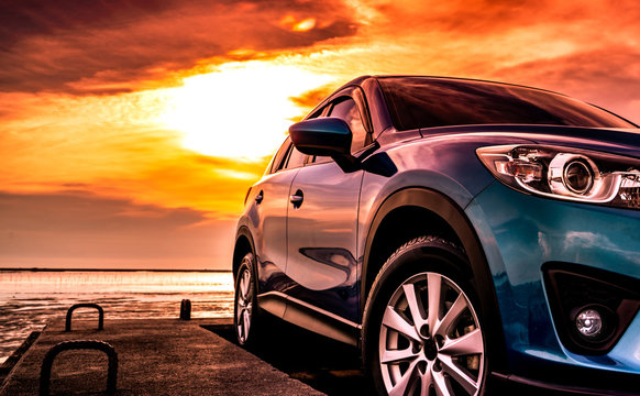 Blue SUV car with sport and modern design parked on concrete road at sunset sea beach with orange sky. Hybrid and electric car technology concept. Automotive industry. Car care service wallpaper.