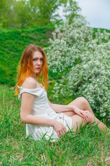 Naklejka na ściany i meble The red-haired girl in a white dress sitting on green grass on a background of a blossoming Apple tree. Girl spring on nature.