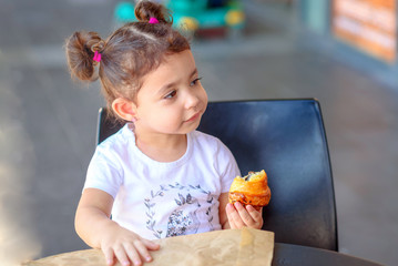 Zero waste concept, paper reusable packaging for grocery shopping. Cute little girl having breakfast in the cafe on the city street. The child is eating a croissant at restaurant , eco paper bag.