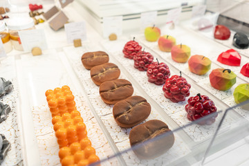 Rows of multicolored cakes and desserts in the window of a pastry shop. Modern mousse dessert, sweet snack. in the form of coffee beans, cherries, apples and hearts.