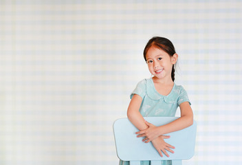 Happy Asian child preschool girl in a Kindergarten room poses on plastic baby chair.