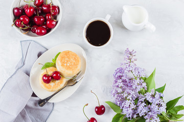 Summer Breakfast with lilac flowers. Cottage cheese pancakes with berry cherry, cup of black coffee, milk and bowl with cherry on white table background. Top view
