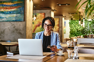 Money is only a tool. Mixed race smiling woman using credit card for shopping and paying bills on-line while sitting in coffee shop