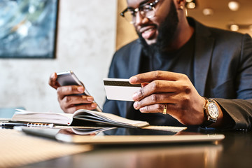 Shot of african american businessman holding mobile and credit card, while resting in the cafe - obrazy, fototapety, plakaty