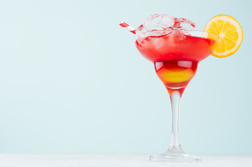 Tropical sunrise striped red, yellow drink with splash, orange, ice cubes, straw in exquisite misted glass on green wall, white wood table.