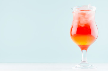 Exotic striped alcohol citrus red and yellow cocktail with  ice cubes in rich wet wineglass on  pastel green background.