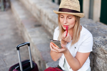 Girl traveler corrects makeup and applies red lipstick outside. Woman tourist with luggage sits at town street in Europe. Concept of student travel, summer vacation, female tourism, trip, adventure.