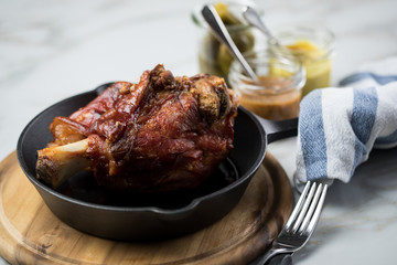 Gegrillte Schweinefleisch Haxe in gusseiserne Pfanne auf Holzbrett mit Cornichons, süßen und mittelscharfen Senf auf Marmor Hintergrund