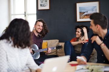 A group of young friends with guitar indoors at home, house sharing concept. - obrazy, fototapety, plakaty
