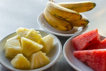 Ripe banana, pineapple and watermelon on white plates on table. Variety of tropical fruits. Exotic breakfast on tropical vacation. Slices of fresh pineapple and red juicy watermelon. Sweet fruits.