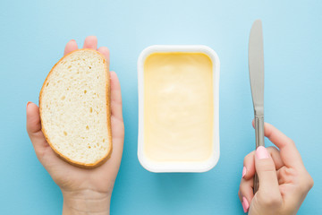 Woman's hands holding slice of white bread and knife. Opened plastic pack of light yellow margarine on pastel blue desk. Preparing breakfast. Point of view shot. Closeup. - obrazy, fototapety, plakaty