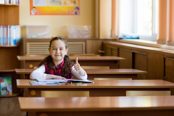 Smiling caucasian girl sitting at desk in class room and showing thumb up gesture. The young pre schoolgirl Happy pupil