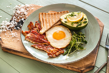 Plate with tasty fried egg, bacon, avocado and toasts on table