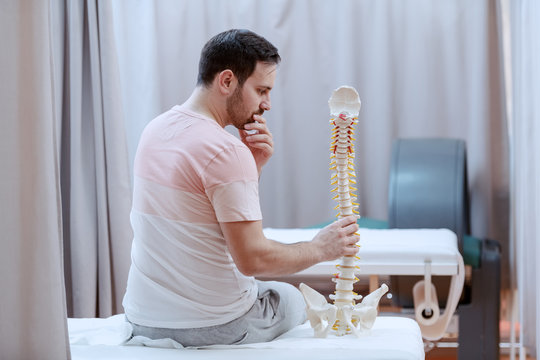 Confused Male Caucasian Patient Holding Spine Model While Sitting On Hospital Bed With Backs Turned.