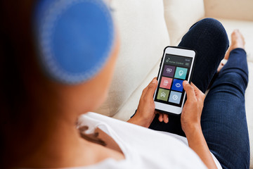 Close-up of young woman in headphones sitting on the couch and using smart home application on mobile phone at home