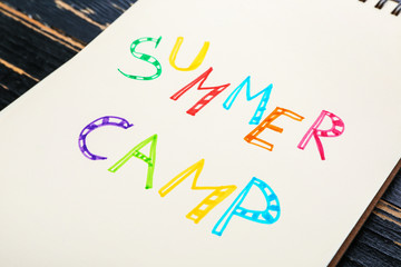 Notebook with words SUMMER CAMP on wooden background, closeup