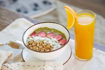 Delicious organic green smoothie bowl garnished with strawberries, sliced almonds and granola served with a glass of fresh squizzed orange juice