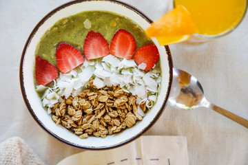 Delicious organic green smoothie bowl garnished with strawberries, sliced almonds and granola served with a glass of fresh squizzed orange juice