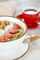 Delicious organic green smoothie bowl garnished with strawberries, sliced almonds and granola served with a cup of coffee