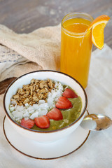 Delicious organic green smoothie bowl garnished with strawberries, sliced almonds and granola served with a glass of fresh squizzed orange juice