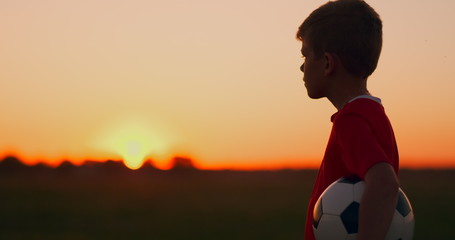 Young football player goes with the ball on the field dreaming of a football career, at sunset looking at the sun
