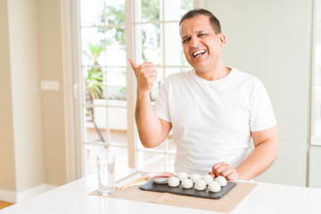 Middle age man eating asian dumplings at home smiling with happy face looking and pointing to the side with thumb up.