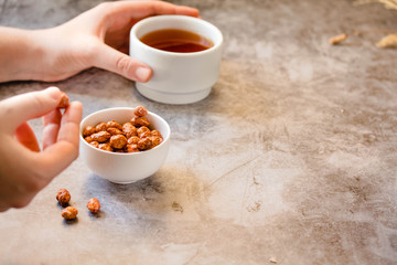 Caramelized peanuts in white bowl and cup of tea on gray background.The process of eating. In the hands of a young woman.Free space for text.