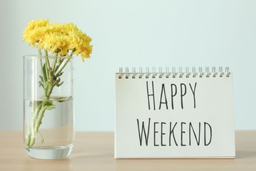 Yellow flower in glass with happy weekend notebook on wood table on white background.