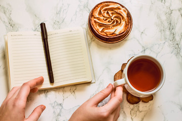 Opened notebook with blank area, pen, laptop, and a cup of black tea on a white marble table in morning time.