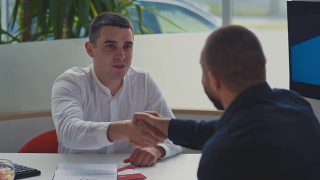 Young salesman giving keys of car to buyer. Men shaking hands in car dealership. Happy buyer