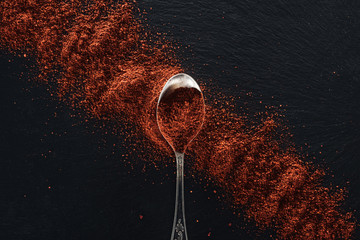 top view of red pepper scattered near silver spoon on textured black background