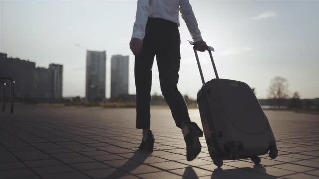 Close up, Knee or ground level shot: Business lady going to taxi parking from the airport or railroad station with her luggage in big sity. Slow motion. Girl in heels stepping and roll suitcase on