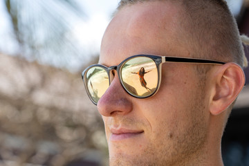Portrait of a young man in sunglasses in which the silhouette of a girl is reflected on the tropical beach near sea