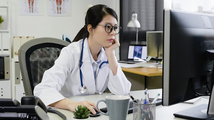 young asian woman doctor working on computer at desk concentrated looking staring on screen wear...