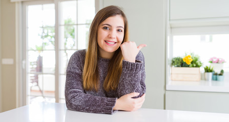 Beautiful young woman at home smiling with happy face looking and pointing to the side with thumb up.